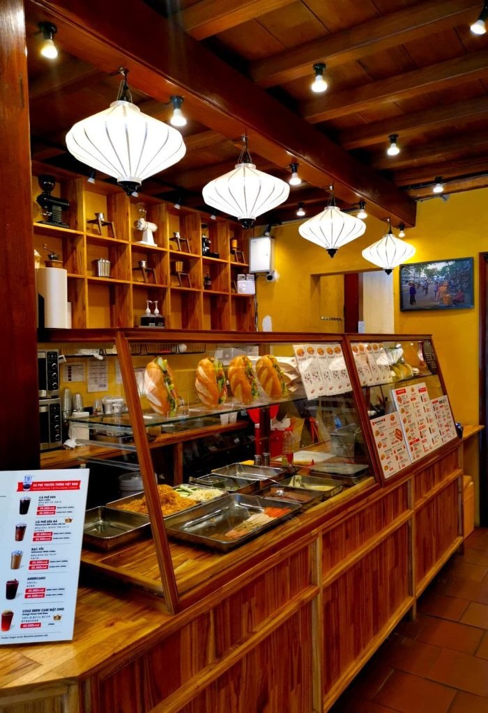 Bên trong quầy gỗ của All Day Banh Mi Hoi An với khay thức ăn và thực đơn trong tủ kính.