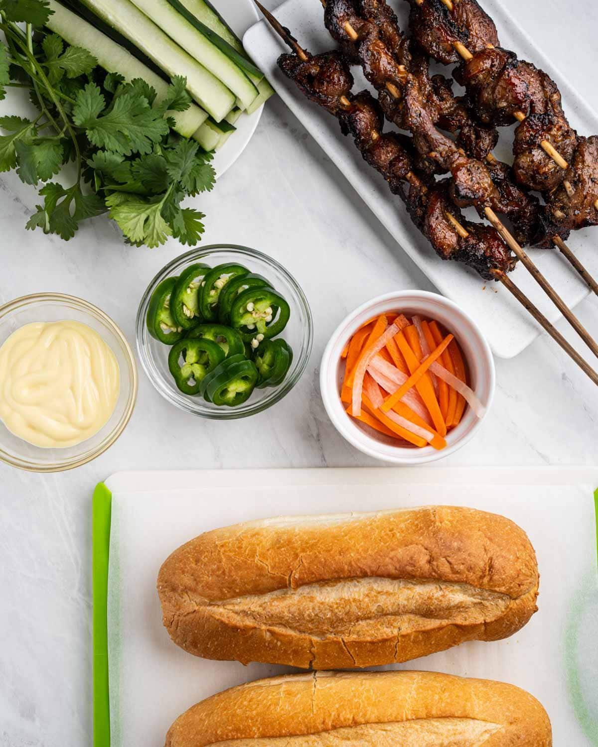 birds eye view of all the ingredients for banh mi thit nuong. Banh mi bread, skewers of grilled pork, pickled carrots and daikon, sliced jalapeños, mayonnaise, cilantro and cucumber.
