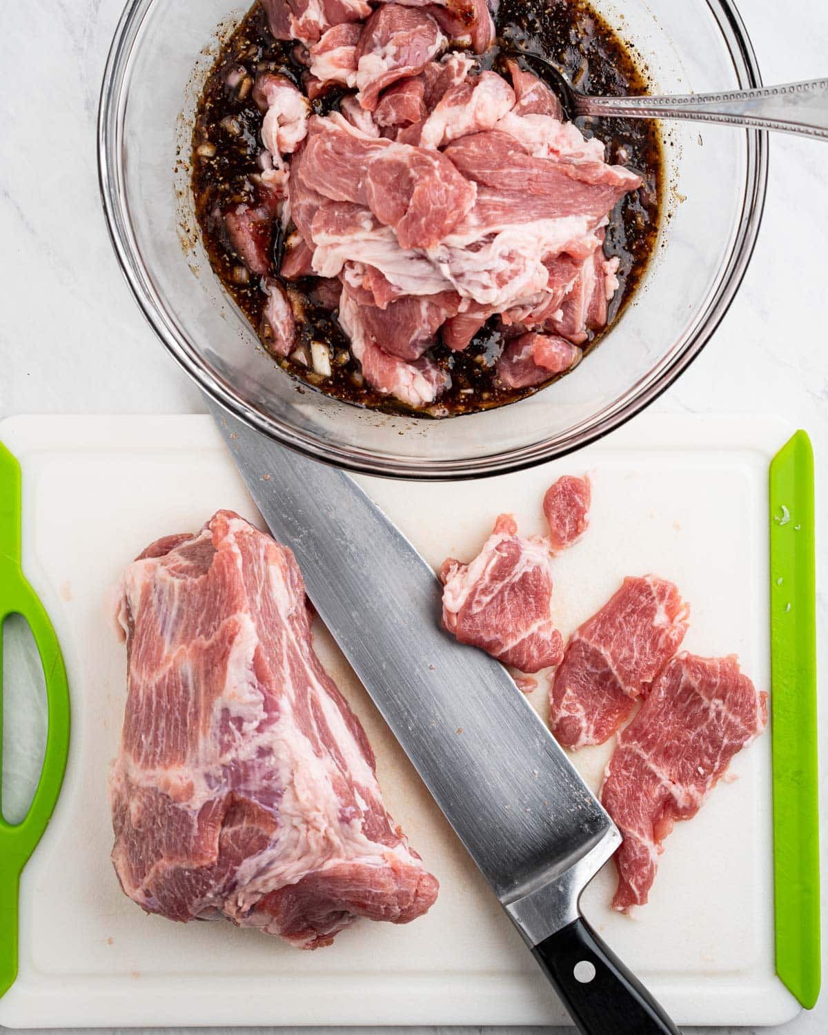 Cutting board with pork, knife thinly slicing pork. Bowl of marinade with sliced pork placed on top.