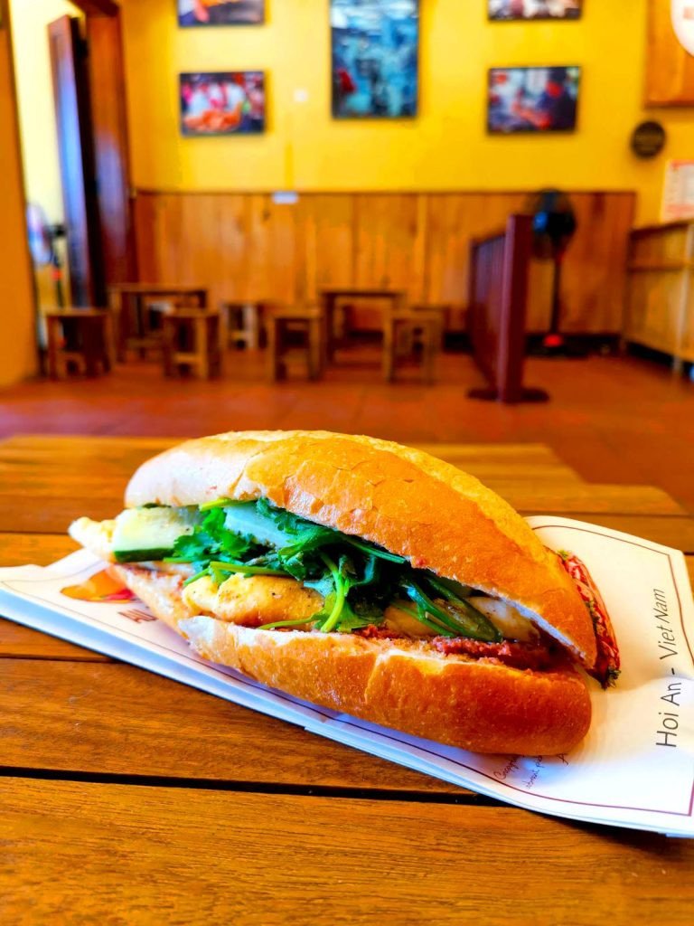 Ổ bánh mì truyền thống với pate, thịt và rau xà lách tươi ngon trên bàn gỗ tại All Day Bánh Mì Hội An